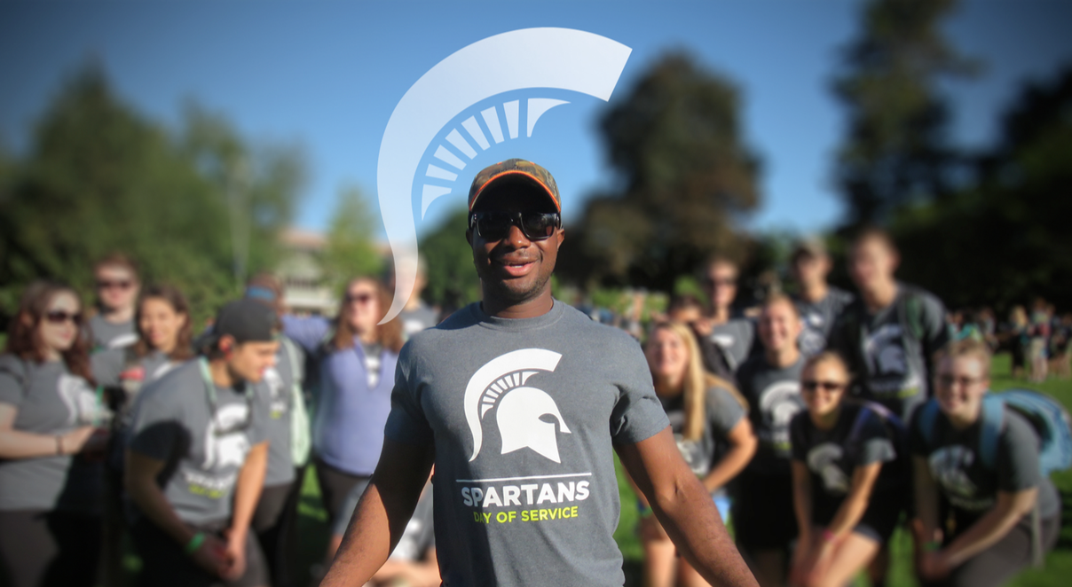 A picture of Spartan Day of Service volunteers. A group of these volunteers stands out of focus in the background.One stands in focus in front of them, wearing a Spartan Day of Service T-shirt and smiling. A graphic of the arc of an MSU Spartan helmet has been edited above the front volunteer's head.